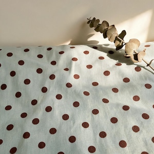 May include: A light mint green fabric featuring a repeating pattern of dark brown polka dots. A single dried eucalyptus branch with rounded leaves rests across the top right corner of the textile, casting a soft shadow onto the surface.
