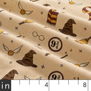 May include: A beige fabric featuring a repeating pattern of brown sorting hats, golden winged balls, striped scarves, round glasses, broomsticks, and the text 9 3/4, with a ruler visible at the bottom showing measurements in inches.