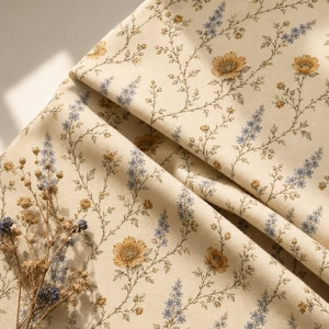May include: A cream-coloured fabric featuring a repeating botanical print of small yellow flowers and light blue sprigs on thin green stems. A small cluster of dried flowers with yellow and blue petals rests on the folded textile surface.