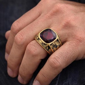 Op de afbeelding: Een gouden ring met een grote, vierkant geslepen, dieppaarse edelsteen in een brede band. De band is versierd met ingewikkelde, donkere, gegraveerde bloemmotieven die contrasteren met het gepolijste gouden oppervlak van de metalen zetting.