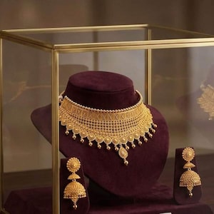 Puede incluir: Un collar tipo gargantilla de oro con intrincados patrones de filigrana y dijes circulares colgantes, junto con pendientes a juego en forma de campana. El conjunto de joyería se exhibe sobre un soporte de terciopelo morado oscuro.