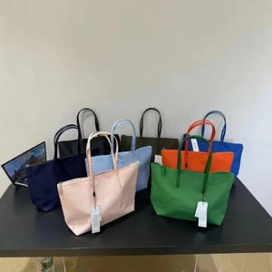 Może przedstawiać: Kolekcja siedmiu prostokątnych toreb typu tote w różnych jednolitych kolorach, w tym czarnym, jasnoniebieskim, jasnoróżowym, oliwkowym, pomarańczowym, jaskrawozielonym i królewskim błękicie, każda z dwoma cienkimi paskami i białą prostokątną metką.