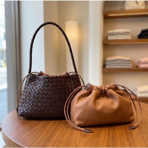 May include: A dark brown woven leather handbag with a braided shoulder strap and a drawstring closure, positioned next to a smaller, smooth tan leather drawstring pouch with matching tan leather cords and a rounded, soft-sided silhouette.
