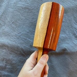 May include: A two-tone wooden mallet, featuring light brown and reddish-brown sections. The mallet is held in a hand, highlighting its contrasting colours and smooth finish. The handle is an extension of the wood grain.