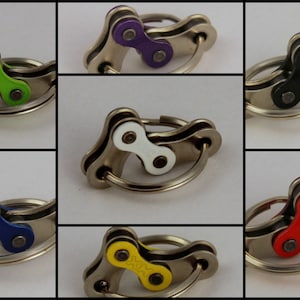 May include: A collection of keychains featuring a bicycle chain link design. Each keychain has a silver-colored ring and a chain link in various colors, including green, purple, black, white, blue, yellow, and red. The keychains are displayed against a neutral background.