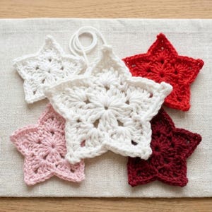 May include: A collection of five star-shaped crochet ornaments in various colors including white, light pink, bright red, and dark red. Each star features a detailed, intricate yarn pattern and one white star includes a small attached hanging loop.