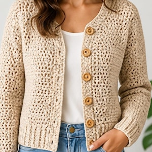 May include: A beige crochet cardigan featuring a round neckline, long sleeves, and a front button closure with five large, round, light brown wooden buttons. The garment is crafted with an open-knit texture and ribbed cuffs at the wrists.