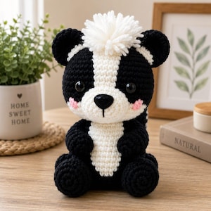 May include: A crocheted skunk plush toy featuring a black and white body, a white pom-pom on top of its head, black safety eyes, a black nose, and small pink stitched cheeks. The skunk is seated in an upright position with a white stripe.