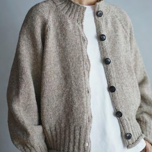 May include: A beige, speckled knit cardigan featuring a ribbed crew neckline, ribbed cuffs, and a ribbed hem. The garment is fastened with six dark, round buttons down the front and is worn over a plain white t-shirt and brown trousers.