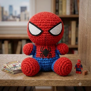 May include: A crocheted Spider-Man plush toy featuring a round red head with large white eyes, a red and blue body with a black spider emblem on the chest, a small Spider-Man minifigure, and a stack of miniature comic books on a wooden shelf.