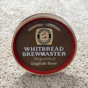 May include: A circular wooden beer tap handle featuring a red outer rim and a central emblem with the text Whitbread Brewmaster. The logo depicts a figure holding a beer mug, flanked by wheat stalks, with the words Imported English Beer below.
