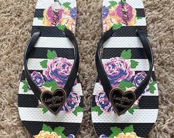 May include: A pair of flip-flops featuring a black and white horizontal striped pattern with a floral print of pink, purple, and yellow roses. Each sandal has a black strap adorned with a gold-rimmed black heart charm labeled Luv Betsey.