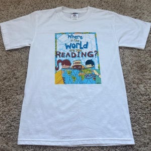 May include: A white short-sleeve t-shirt featuring a graphic print of three cartoon characters looking at a world map. The text reads, Where in the WORLD are you READING? and includes the word ATLAS on the map. The design uses blue, yellow, and brown.