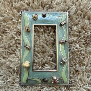 May include: A rectangular light switch wall plate featuring a marbled sage green and light green enamel finish. The frame is decorated with small metal charms including a heart, a baby shoe, a spool, a duck, a rattle, and a teddy bear.