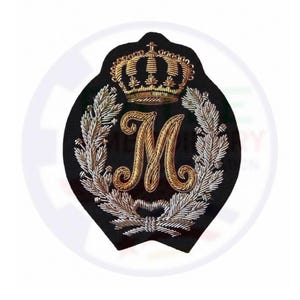 May include: A black embroidered patch featuring a central gold letter M, topped with a detailed gold crown. The letter is surrounded by a silver laurel wreath design, with the entire emblem stitched onto a dark fabric base in a shield shape.