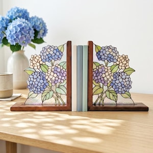 May include: A pair of L-shaped wooden bookends featuring stained glass panels with a floral hydrangea design in shades of blue, purple, and cream with green leaves. The bookends hold two books upright between them on a flat wooden surface.
