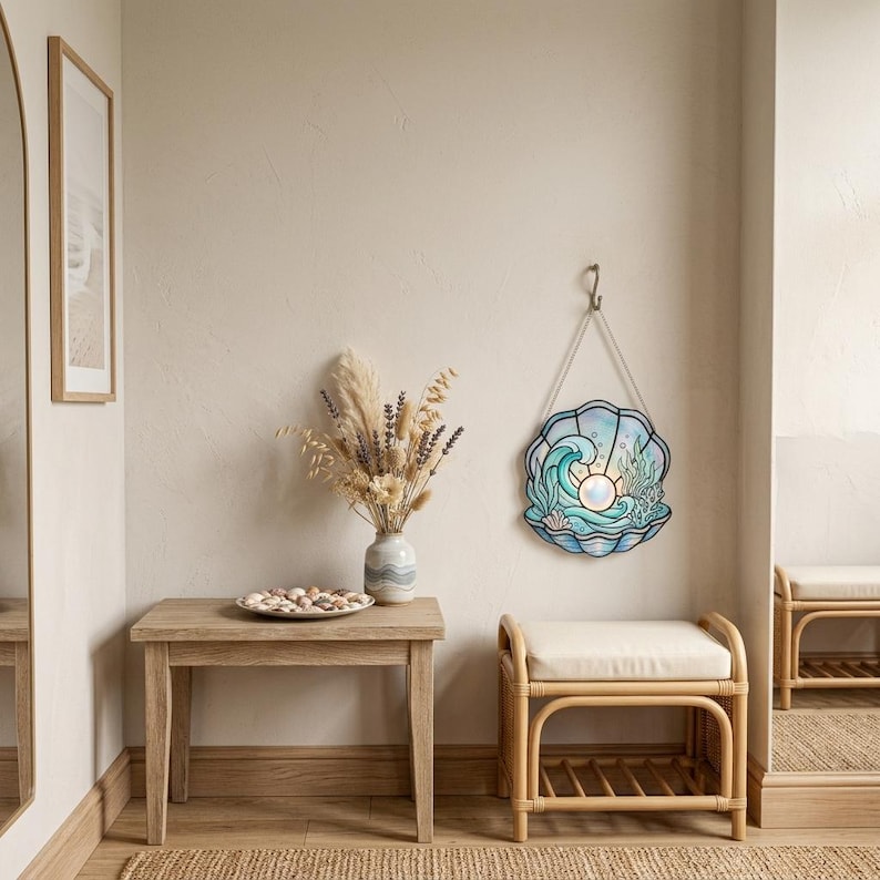 Puede incluir: Un adorno de pared de vitral con forma de concha de abanico, que presenta una perla central brillante, olas azules y algas verdes. La pieza est&aacute; suspendida por un cord&oacute;n fino de un gancho met&aacute;lico, mostrando detalles intrincados de plomo.