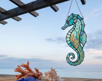 Puede incluir: Un atrapasoles de vidrieras con forma de caballito de mar en tonos azul, verde y turquesa con un diseño de coral naranja, suspendido por una cadena plateada. El caballito de mar tiene una pequeña cuenta blanca como ojo y líneas de soldadura.