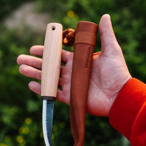 May include: A fixed-blade knife with a light-colored wooden handle and a dark, textured metal blade, accompanied by a brown leather sheath featuring the embossed text "BERSAKNIVES" on the front surface, designed for carrying the tool.