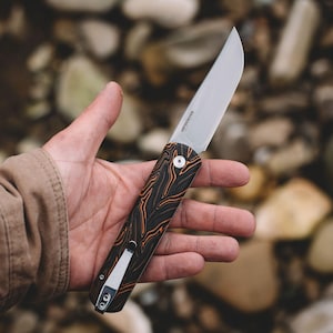 May include: A folding pocket knife with a silver metal blade and a black handle featuring orange marbled patterns. The handle includes a silver metal pocket clip attached to the bottom. The blade has the text KIZER KNIVES printed on the side.