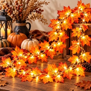May include: A string of artificial maple leaf lights featuring vibrant orange, red, and yellow fabric leaves illuminated by small warm white LED bulbs. The flexible wire strand is draped across a wooden surface, creating a glowing autumn display.