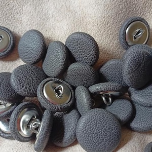 Op de afbeelding: Een verzameling ronde knopen bekleed met een donkergrijs, getextureerd kunstlederen materiaal. Elke knoop is voorzien van een gladde, zilverkleurige metalen schacht aan de achterzijde, ontworpen voor bevestiging aan kleding of stoffen projecten.