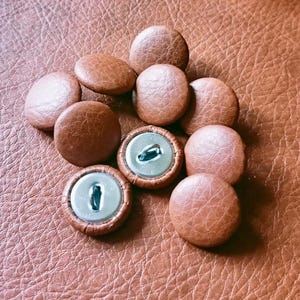 May include: A collection of round buttons covered in a textured brown material. Each button features a smooth, domed top surface with a visible grain pattern. Two of the buttons are flipped over, revealing a silver-toned metal back with a central loop.