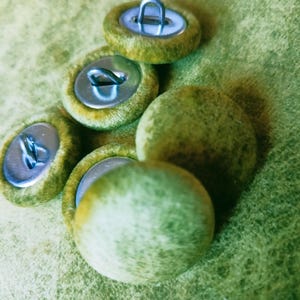 Op de afbeelding: Een verzameling van vijf ronde knopen bekleed met een textuurrijke, mosgroene stof. Elke knoop is voorzien van een zilverkleurig metalen oogje aan de achterzijde. Het stoffen oppervlak vertoont een gevlekt, groen patroon.