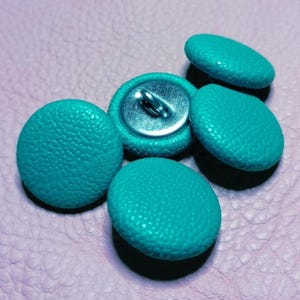 May include: A collection of four round buttons covered in a textured, bright turquoise fabric. Each button features a circular, pebbled surface finish and a metallic silver-toned shank on the underside for attachment to garments or craft projects.
