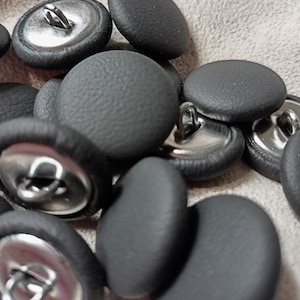 May include: A collection of round buttons featuring a textured dark gray surface. Each button has a smooth, rounded top and a metallic silver-toned back with a sturdy loop shank for attachment, creating a uniform appearance across the set.