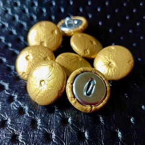 Op de afbeelding: Een verzameling ronde, goudkleurige knopen met een getextureerd oppervlak. Elke knoop heeft een centrale inkeping en een metalen achterkant met een lus voor bevestiging. De knopen zijn in een groep gerangschikt en tonen zowel de voor- als achterkant.