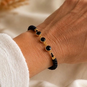 May include: A bracelet featuring three round, black faceted stones set in gold-toned metal bezels, connected by a thin gold-toned chain, and finished with a black beaded cord band that wraps around the wrist for a secure and adjustable fit.
