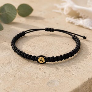 Op de afbeelding: Een zwart koordarmbandje met een centrale ronde goudkleurige bedel waarin de letter A in het zwart is gegraveerd. De armband is gemaakt met een gevlochten knooppatroon en heeft verstelbare trekkoorden aan de achterkant voor een goede pasvorm.