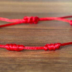 May include: A bright red cord bracelet featuring a series of evenly spaced, hand-tied knots along the length of the string. The bracelet includes an adjustable sliding knot closure mechanism with two loose cord ends extending from the center.