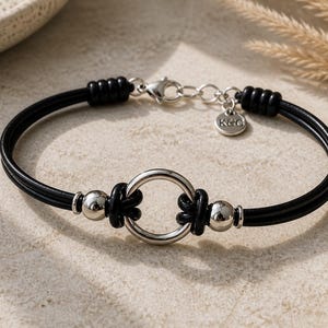 Op de afbeelding: Een zwart koordarmband met een centrale zilverkleurige metalen ring geflankeerd door twee zilverkleurige kralen. De armband is voorzien van een karabijnsluiting, een verstelketting en een kleine ronde bedel met de letters K&C.