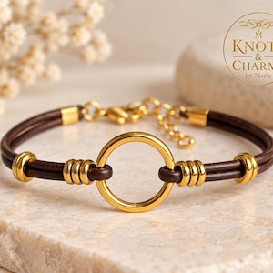 May include: A dark brown leather cord bracelet featuring a central gold-toned metal ring, accented by smaller gold-toned metal beads along the cord. The bracelet includes a gold-toned metal chain extension and a lobster clasp closure.