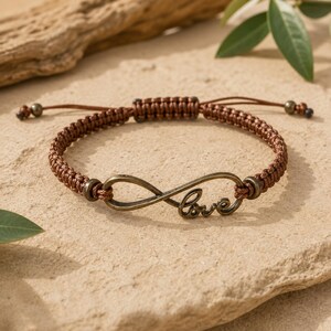 May include: A brown braided cord bracelet featuring a metallic infinity symbol charm with the word love integrated into the design. The bracelet includes a sliding knot closure and small metallic beads at the ends of the adjustable cords.