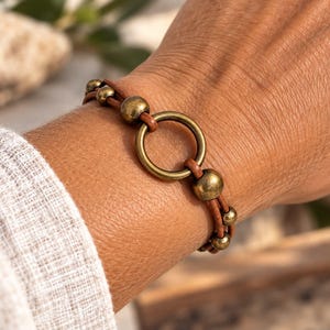 Op de afbeelding: Een armband van bruin leren koord met een centrale ring van messingkleurig metaal, geflankeerd door verschillende gladde, bolvormige messingkleurige kralen. De armband is opgebouwd uit twee leren koorden die de metalen onderdelen vasthouden.