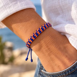 Könnte beinhalten: Ein geflochtenes Kordelarmband mit abwechselnden leuchtend blauen und hellen orangefarbenen Strängen. Das Armband hat ein gewebtes, geknotetes Design und zwei herabhängende blaue Kordelenden mit Knoten an den Spitzen.