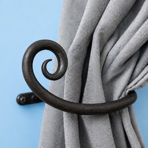 Puede incluir: Un alzapaños de cortina de metal marrón oscuro con un extremo curvo en forma de espiral. El herraje está montado en una pared y sujeta una cortina de tela gris recogida. El metal tiene un acabado mate texturizado con tornillos visibles.