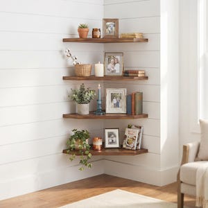 Puede incluir: Cuatro estantes esquineros de madera marr&oacute;n oscuro montados en una pared blanca con paneles. Los estantes sostienen varios art&iacute;culos decorativos, incluyendo plantas en macetas, marcos de fotos, velas, libros y una cesta tejida en una disposici&oacute;n.