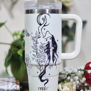 May include: A white stainless steel tumbler with a handle and a clear lid, featuring a black line art illustration of a figure reaching toward a crescent moon, surrounded by roses, leaves, stars, and a butterfly. The brand name TYESO is printed.