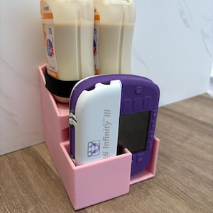 Puede incluir: Un organizador de plástico rosa claro de varios niveles que contiene dos botellas de suplemento nutricional beige de 500 ml y un dispositivo médico Infinity III morado y blanco con pantalla y botones para organizar suministros médicos.