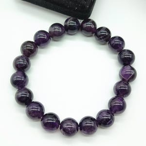 May include: A circular bracelet composed of polished, spherical amethyst beads in deep shades of purple. The beads are strung together on an elastic cord, showcasing natural variations in color and translucency within each individual stone.