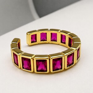 Pode incluir: Um anel aberto em tom dourado com uma série de pedras retangulares em rosa magenta vibrante dispostas numa fila contínua. A banda é construída com molduras metálicas individuais para cada pedra, criando um aspeto geométrico estruturado.