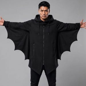 Op de afbeelding: Een zwarte hoodie met ritssluiting en geïntegreerde vleermuisvleugelpanelen die van de mouwen tot aan de romp lopen. Het kledingstuk is voorzien van een capuchon met trekkoord, lange mouwen met geribbelde boorden en twee verticale zakken met rits.