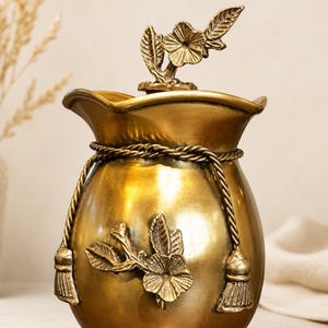 May include: A metallic gold-toned jar with a rounded body and a flared, wavy rim. The jar features a decorative rope cord tied around the neck with two tassels, and a sculpted floral branch motif on the front. A matching lid has a floral handle.