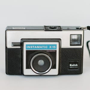 May include: A vintage Kodak Instamatic X-15 camera. The camera has a black and silver body with a blue and white front panel. The camera has a black strap and the text "Instamatic X-15 Camera" is visible.