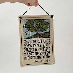 May include: A rectangular ceramic wall hanging with a black cord for hanging. The artwork features a tree design with a blue background and the quote "Promise me you'll always remember..." by Christopher Robin.
