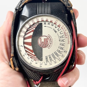 May include: Vintage DeJur exposure meter in a black leather case. The meter has a circular dial with numbers and markings in black, red, and white. The case is worn, showing signs of age. A red cord is attached to the meter.